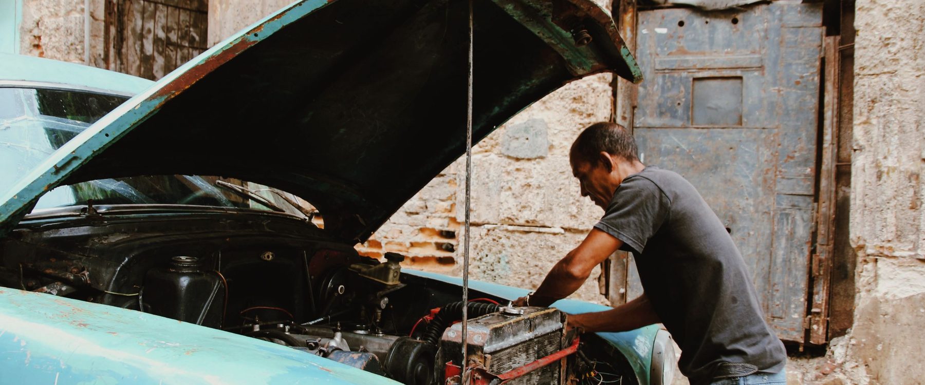 Mecânico debruçado sobre o motor de um carro antigo de capô aberto, diante de uma parede de pedra e de uma porta de ferro enferrujada